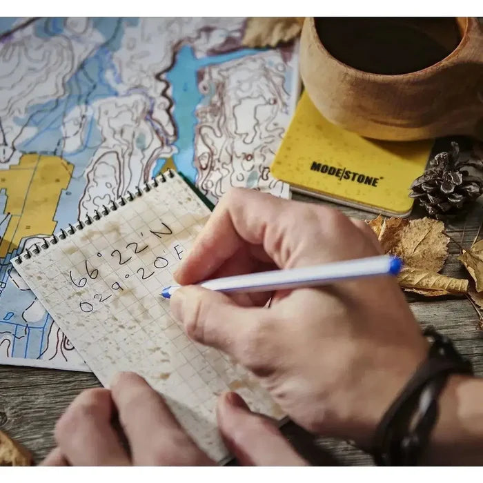 Person using the Modestone A6 Top Spiral Waterproof Notepad outdoors, writing coordinates on waterproof grid paper beside a map and coffee cup.  from  John Bull Clothing