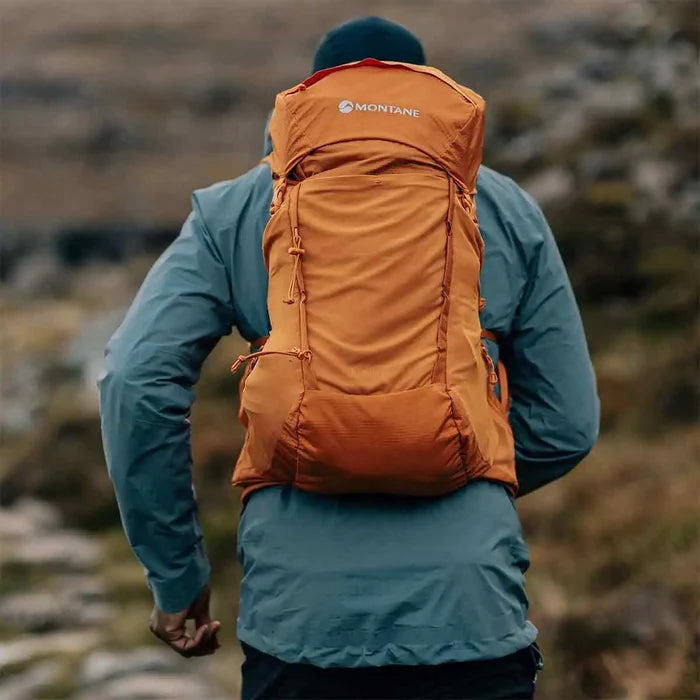 Hiker wearing the Montane Trailblazer 25L Rucksack in orange, trekking through rugged outdoor terrain with a secure fit.  from  John Bull Clothing