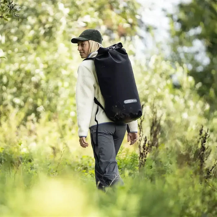 Woman hiking through forest with Ortlieb X-Tremer 35L Carry Bag Black on her back, demonstrating its suitability for outdoor exploration and travel.  from  John Bull Clothing