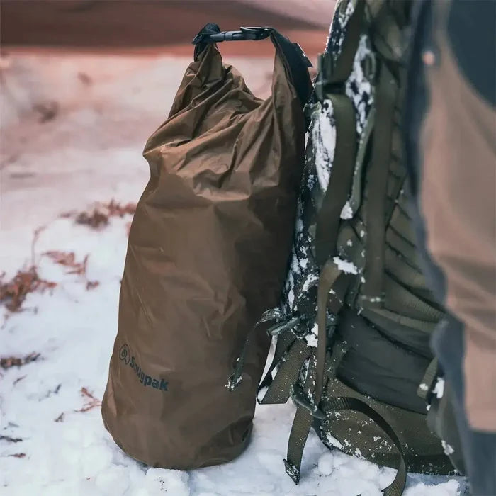 Snugpak Dri-Sak Waterproof Dry Bag in brown, attached to a backpack in a snowy outdoor setting, ensuring dry storage in harsh conditions.  from  John Bull Clothing