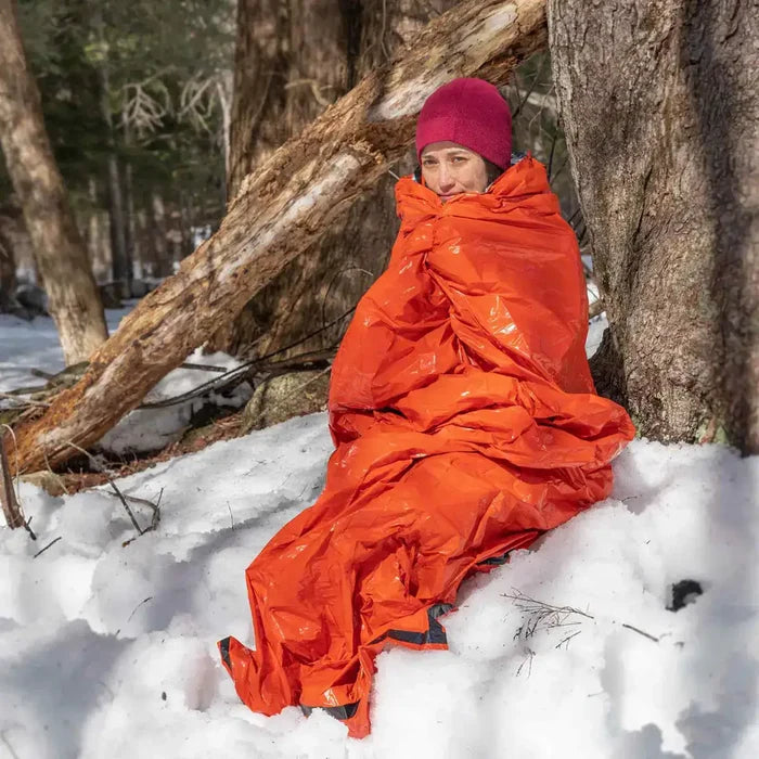 Person wrapped in SOL Emergency Bivvy Bag Shelter with Whistle, sitting in snow for cold-weather survival protection.  from  John Bull Clothing