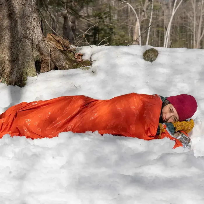 Camper sleeping in SOL Emergency Bivvy Bag Shelter with Whistle on snow, demonstrating insulation and emergency use.  from  John Bull Clothing