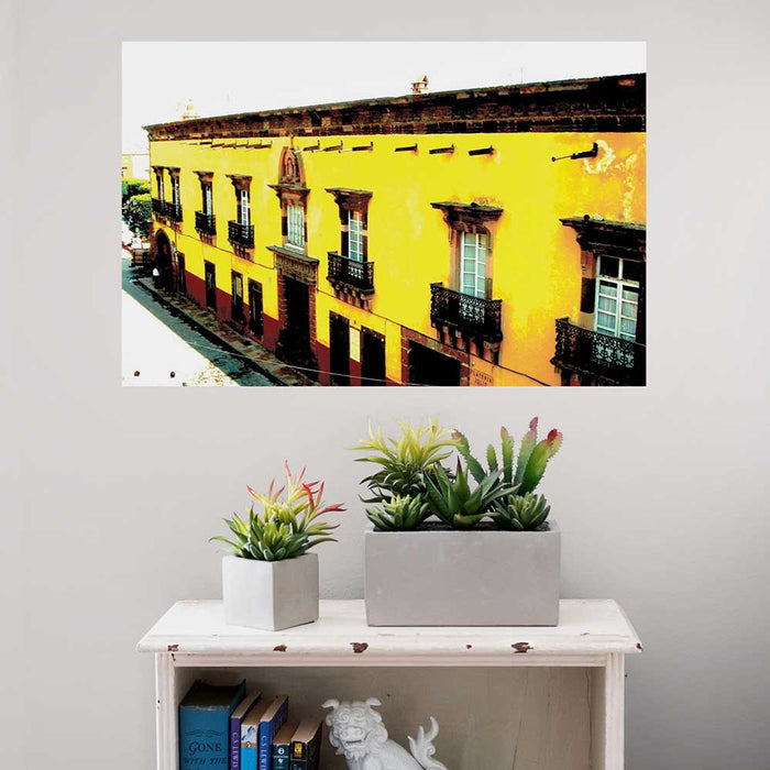 24 inch decal of yellow Mexican hotel with balconies installed on a wall above a white shelf with plants and books.