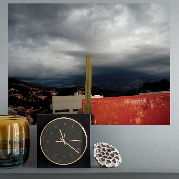 24 inch poster of a dark stormy sky in Mexico with potted cactus in foreground displayed on a wall above clock and vase